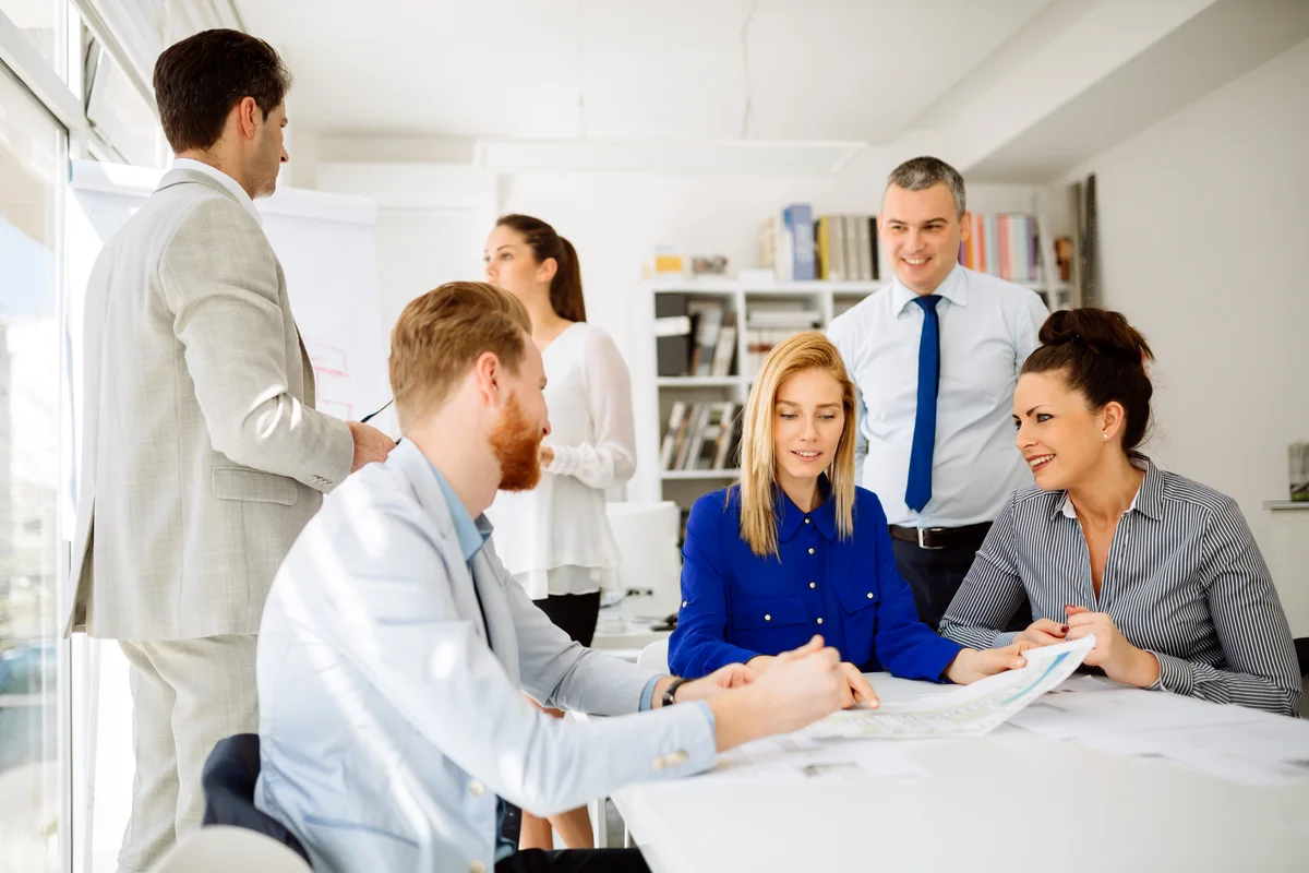 A business group meets around a table while one colleague presents.